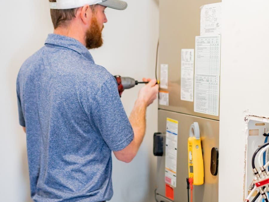 HVAC technician in a blue polo and cap drilling or assembling a furnace panel in a utility room with a yellow meter attached nearby, cables visible on the wall